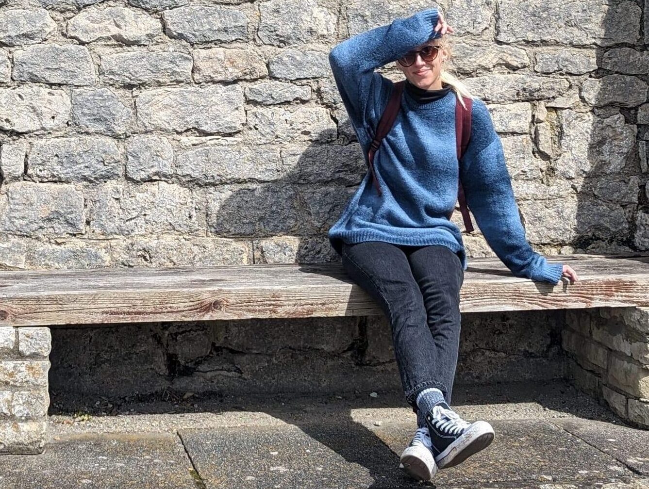 Lune sitting on a bench in a blue sweater, shielding their eyes from the sun with their arm.